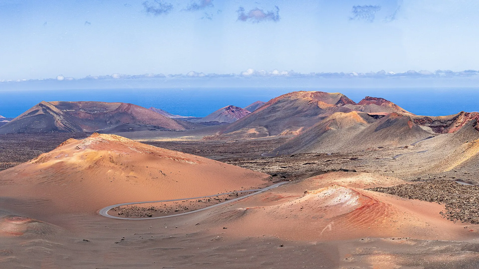 Rote und ockerfarbene Vulkane im Nationalpark Timanfaya, dazwischen eine gewundene Straße und dahinter das Meer